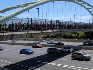 Around a hundred people stand on a pedestrian overpass over the highway, holding flags and signs